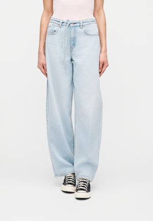 Isik, kes kannab helesiniseid laiade säärega teksapükse kangavööga, mis on vöökohal seotud, ning musti kõrgema säärega tenniseid valgete paeltega.