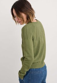 Pull en tricot vert olive avec un yoke texturé et un ourlet côtelé, associé à un jean en denim bleu. Le tissu semble doux et chaud.