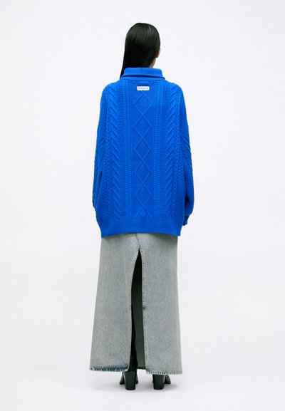 Suéter de punto azul oversized con patrones de trenzado y un cuello alto, combinado con una falda larga de mezclilla gris que presenta una abertura en la parte trasera.