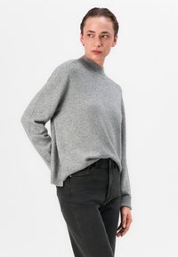 Grau pullover mit hohem Kragen, gerippten Bündchen und Saum. Wird mit einer lockeren Passform über schwarzen Jeans getragen, die eine glatte Textur aufweisen.