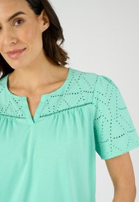 Top verde menta con scollo a V, caratterizzato da un intricato motivo a taglio laser sulle maniche e sul giogo. Tessuto morbido con vestibilità rilassata.