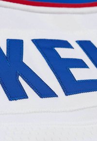 Weißer Stoff mit großen, blauen gestickten Buchstaben und einer strukturierten Oberfläche. Rote und blaue Akzente sind an den Rändern sichtbar.
