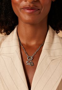 Zilverkleurige ketting met twee speelse hangers in de vorm van de cijfers '2' en '0', gedragen bij een lichtbeige, getextureerde blouse met kraag.