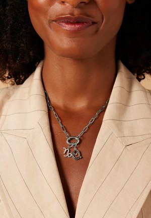 Zilverkleurige ketting met twee speelse hangers in de vorm van de cijfers '2' en '0', gedragen bij een lichtbeige, getextureerde blouse met kraag.