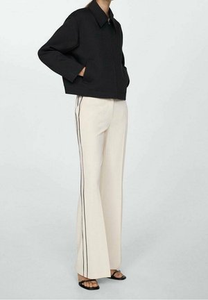 Personne portant une veste courte noire avec des poches latérales, un pantalon large crème avec des bandes noires sur les côtés et des sandales noires à bout ouvert.