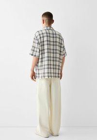 Lyhythihainen, kauluksellinen paita beigen ja mustan ruudullisen kuosin kanssa, yhdistettynä leveisiin kermaisiin housuihin ja vaaleisiin lenkkareihin.