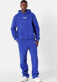 Conjunto de sudadera con capucha y pantalones de chándal en color azul, fabricado en un tejido suave, con un bolsillo tipo canguro y un logo en el pecho y el muslo en blanco.