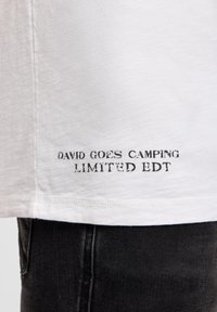 White textured shirt hem with black faded text "DAVID GOES CAMPING LIMITED EDT" worn over black denim jeans.