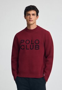 Roter Rundhals-Pullover aus strukturiertem Strickstoff, mit einem schwarzen "POLO CLUB"-Logo in der Mitte. Rippbündchen an den Ärmeln und am Saum.