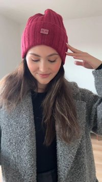 Punane soonikkoeline kootud müts väikese brändisildiga, kantud tekstuurse halli mantli ja musta topi kohal. Pikk pruun juuksed osaliselt nähtavad.