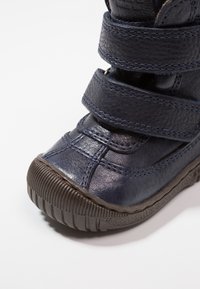 Marineblauwe leren laars met twee verstelbare klittenbandriemen, een gestructureerd oppervlak en een rubberen zool met horizontale groeven voor grip.