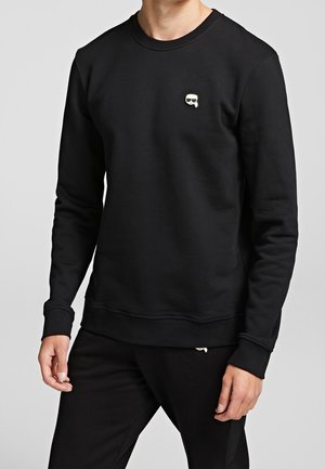 Man draagt een zwarte longsleeve-trui met een klein wit logo van een gezicht met een zonnebril op de linkerborst.