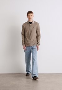 Camisa polo de mangas longas castanha com colarinho em contraste, combinada com calças de ganga largas azul claro e sapatos castanhos. Conjunto simples e casual.
