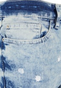 Des jeans en denim bleu clair présentent une texture délavée, une poche arrière avec des détails de couture, et des éclaboussures de peinture blanche pour un look usé.