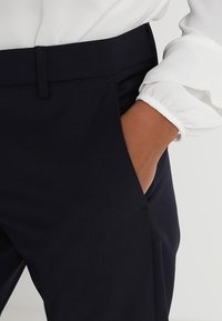 Marinblå skräddarsydda byxor med en slät textur, som har sidofickor och ett rent midjeband. Tyget verkar vara strukturerat och hållbart.