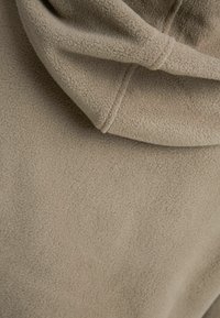 Beige fleece-kangas, jossa pehmeä rakenne, hupulla ja näkyvillä ompeleiden yksityiskohdilla. Materiaali vaikuttaa lämpimältä ja mukavalta.