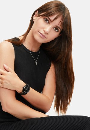 Schwarze Armbanduhr mit rechteckigem Ziffernblatt, silbernen Ziffern und einem strukturierten Armband, getragen von einer Person in einem schwarzen, gerippten, ärmellosen Oberteil.