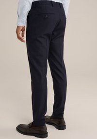 Marineblauwe nauwsluitende broek met een gestructureerd weefsel, riemlussen en twee achterzakken met knopen. In combinatie met een paar bruine instappers.