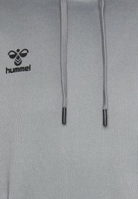 Sweatshirt à capuche en tissu gris clair avec une surface texturée. Équipé d'un logo noir et de cordons de serrage avec des embouts noirs.