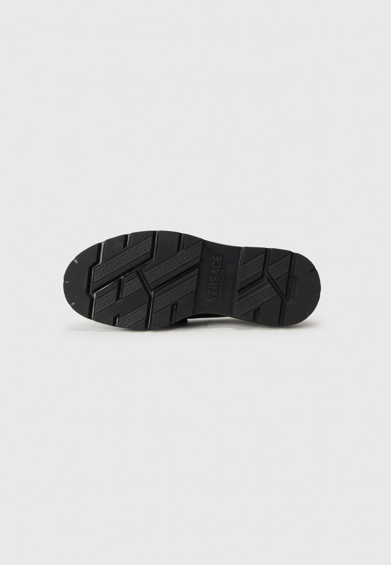 Semelle de chaussure en caoutchouc noir avec un motif de relief texturé, présentant des lignes surélevées et un branding. Construction durable pour une adhérence et une traction.