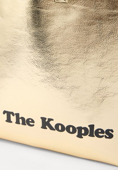 Zlatna umjetna kožna torba koja ima teksturiranu površinu, s imenom brenda "The Kooples" ispisanim debelim crnim slovima duž donjeg dijela.