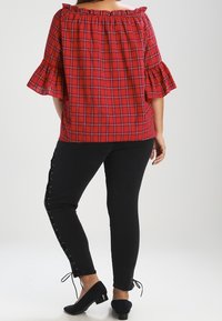 Haut à carreaux rouge dénudé d'une épaule avec des manches volantées, associé à un pantalon noir avec des détails de lacets sur les côtés et des ballerines noires.