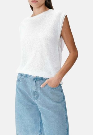 Femme portant un haut blanc texturé sans manches et un short en jean bleu clair mi-genou, main dans la poche, sur fond uni.