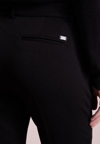 Schwarze, enganliegende Hose mit glatter Textur, ausgestattet mit einer Reißverschluss-Gesäßtasche und einem kleinen silbernen Logo-Detail auf der Tasche.