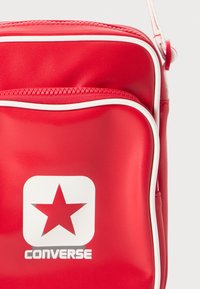 Borsa a tracolla sintetica rossa con accenti bianchi, dotata di una tasca con zip e di un grande logo a stella bianca sopra il nome del marchio "Converse."