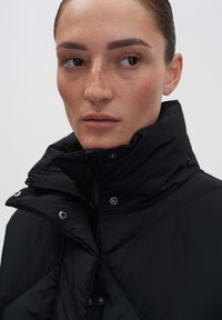 Schwarze Daunenjacke mit hohem Kragen, ausgestattet mit einem Frontreißverschluss und Druckknöpfen. Der Stoff hat eine glatte, gesteppten Textur.