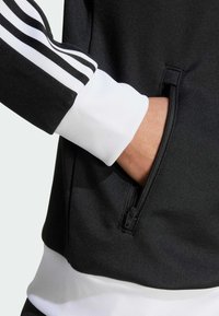 Svart sweatshirt med vita accenter. Har tre svarta ränder på ärmen, en ficka med dragkedja och ribbade vita muddarna vid handlederna.