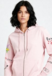 Roza dukserica na patent od mekog materijala. Ima prednje džepove, kapuljaču sa vezicama te dekorativne otiske cvijeća i teksta na rukavima.