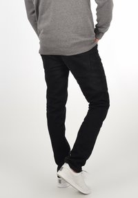 Jeans ajustados negros con una textura suave, con bolsillos traseros y combinados con zapatillas blancas. El fondo es neutro y poco intrusivo.