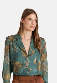 Blusa azul verdoso con mangas largas, patrones florales en oro y verde, escote en V profundo y cierre de botones, con textura de tela transparente.