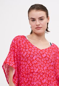 Giovane donna con i capelli tirati indietro che indossa un abito floreale rosso con fiori rosa e la parola "SUN" in testo rosa su uno sfondo bianco.