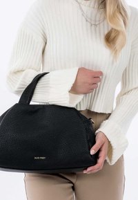 Sac à main en cuir noir granuleux avec une forme courbée, deux poignées courtes et un logo doré embossé. Porté avec un pull blanc côtelé.