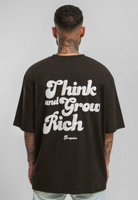 Pruun oversized t-särk, mille seljal on suur valge tekst "Mõtle ja kasva rikkaks" ning brändi logo hemme lähedal.