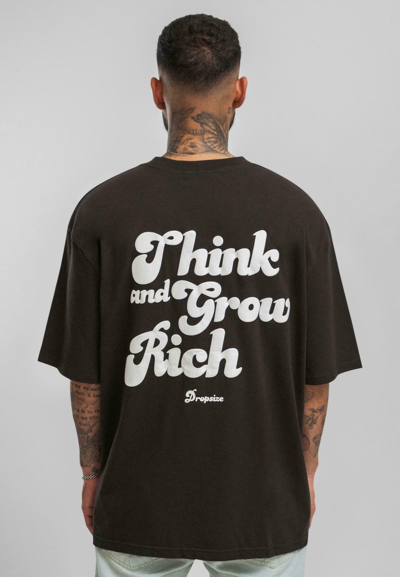 Pruun oversized t-särk, mille seljal on suur valge tekst "Mõtle ja kasva rikkaks" ning brändi logo hemme lähedal.