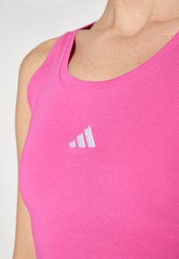 Rózsaszín, testhez simuló ujjatlan felső bordázott anyagból, lekerekített nyakkivágással. A bal oldalon egy kis fehér Adidas logó található.