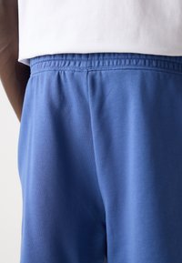 Pantaloni de trening din bumbac albastru, cu talie elastică, având o textură fină și detalii de cusătură subtile, parțial vizibili sub un top alb.