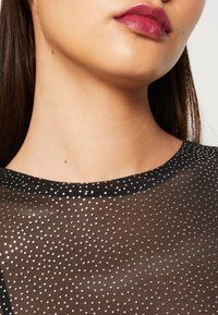 Svart genomskinlig topp med silverprickar, rund urringning och subtilt texturerad tyg. Närbild av urringningen och hakan.