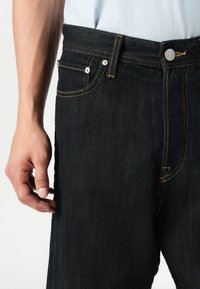 Jeans de mezclilla oscura con costuras amarillas, que presentan bolsillos frontales y un cierre de botón metálico. La tela tiene una textura suave.