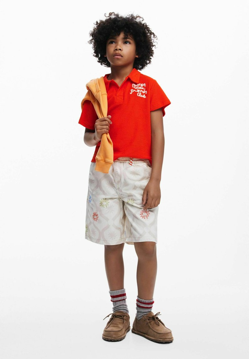 Czerwona koszulka polo z krótkim rękawem z białym haftem, białe szorty w wzory z kolorowymi kwiatami, beżowe buty i szare-siniakowe skarpetki w paski.