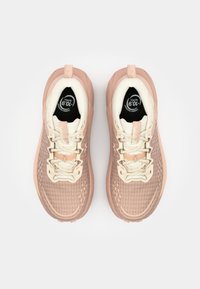 Zapatillas de deporte rosa claro con parte superior de malla texturizada, detalles en crema y elementos reflectantes. Cuerdas planas y suela de goma duradera.