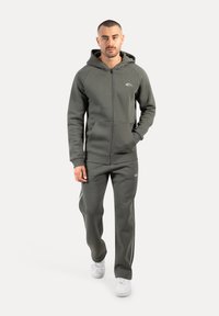 Grauer Zip-Hoodie mit Fronttaschen und Kapuze, kombiniert mit passenden Hosen, die weiße Seitenstreifen und weiße Sneakers haben.