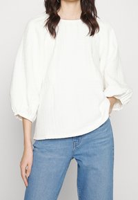 Vit strukturerad topp med rund halsringning och puffärmar, i kombination med blå denimjeans. Tyget har ett rutigt mönster.