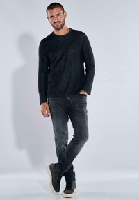Schwarzes Langarmshirt, weiches Material, lässige Passform. Trägt dazu dunkelgraue Jeans und graue High-Top-Sneaker, entspannte Haltung, neutraler Hintergrund.