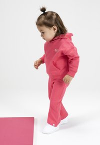 Roze hoodie en broek set van zacht materiaal, met een voorzak en logo. Gecombineerd met witte sneakers, kind in een speelse houding.