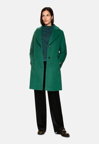 Grüner Wollmischmantel mit Reverskragen, zwei Knöpfen, Seitentaschen, getragen über einem strukturierten blauen Pullover und schwarzen weitgeschnittenen Hosen.