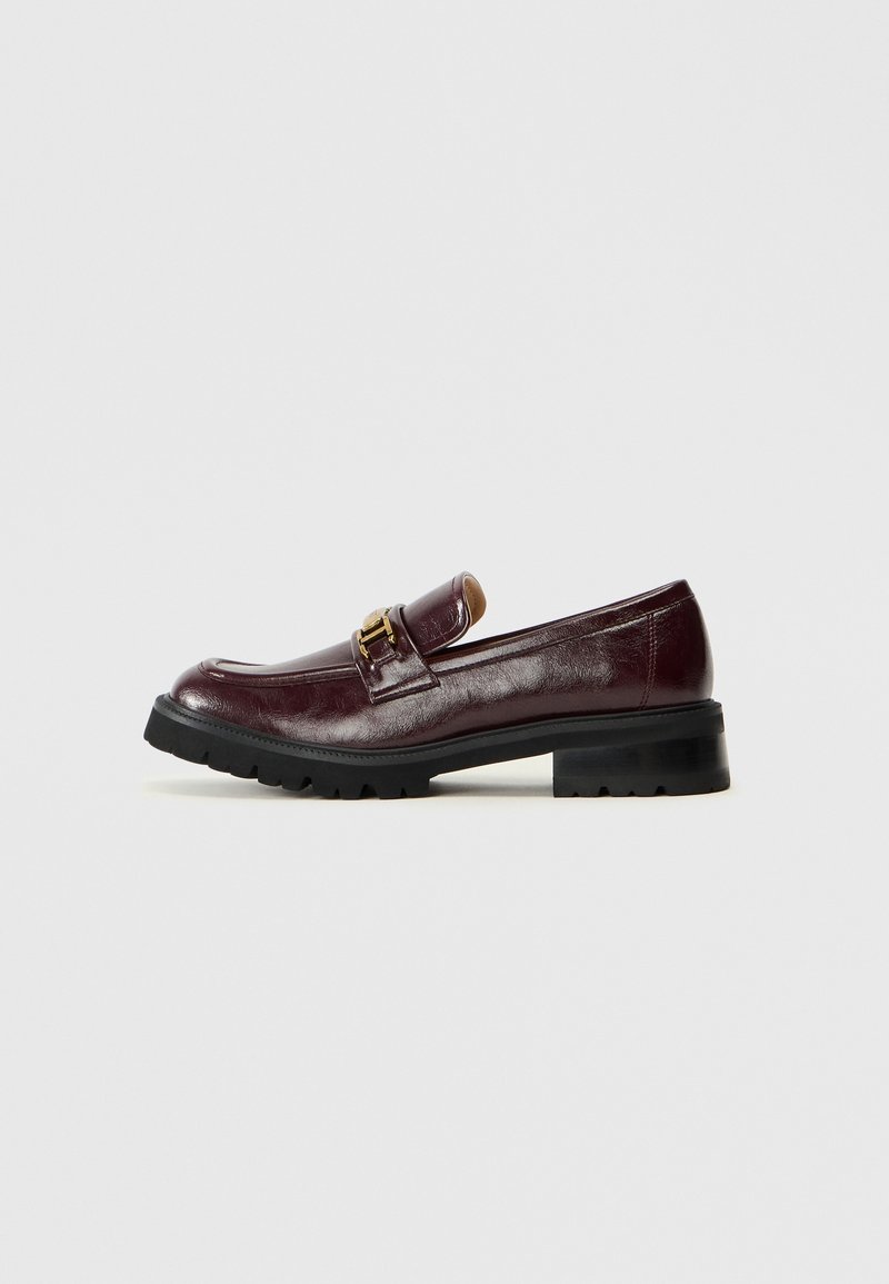 Mocassin en cuir marron foncé avec une semelle épaisse noire et une boucle métallique dorée sur un fond clair uni.